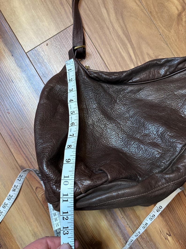 May include: A brown leather crossbody bag with a long adjustable strap. The bag is measured at approximately 13 inches in height. The bag has a zipper closure and a rounded shape. The bag is made of a textured leather material.