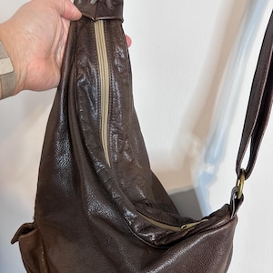 May include: A brown leather sling bag with a zipper closure and adjustable strap. The bag has a textured surface and a drawstring detail at the top. The strap is attached with a gold-colored ring.