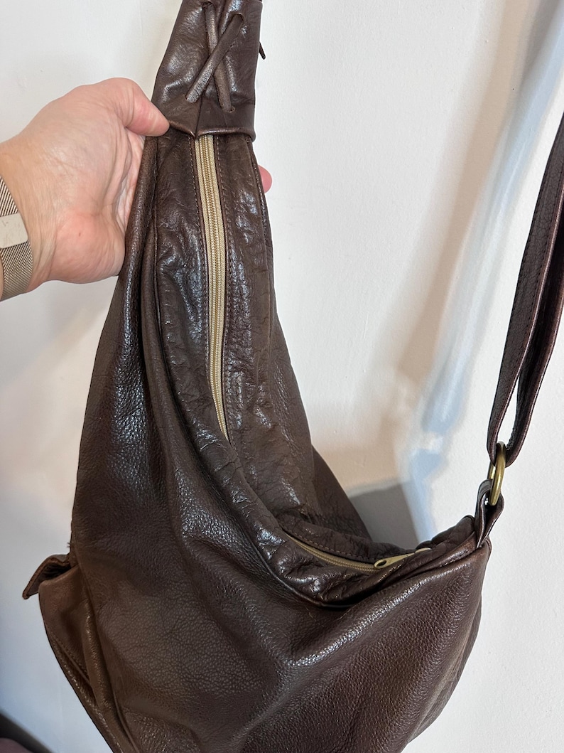 May include: A brown leather sling bag with a zipper closure and adjustable strap. The bag has a textured surface and a drawstring detail at the top. The strap is attached with a gold-colored ring.