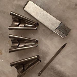 May include: An assortment of metal tools, including three silver-colored, curved clips, a rectangular silver box, and a thin, flat metal piece. The tools are arranged on a light-colored surface.