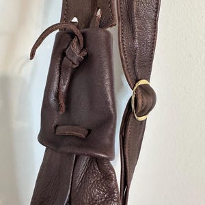 May include: Close-up of a dark brown leather bag with a drawstring closure. The bag features a long strap with a gold-colored metal ring. The leather has a textured appearance.