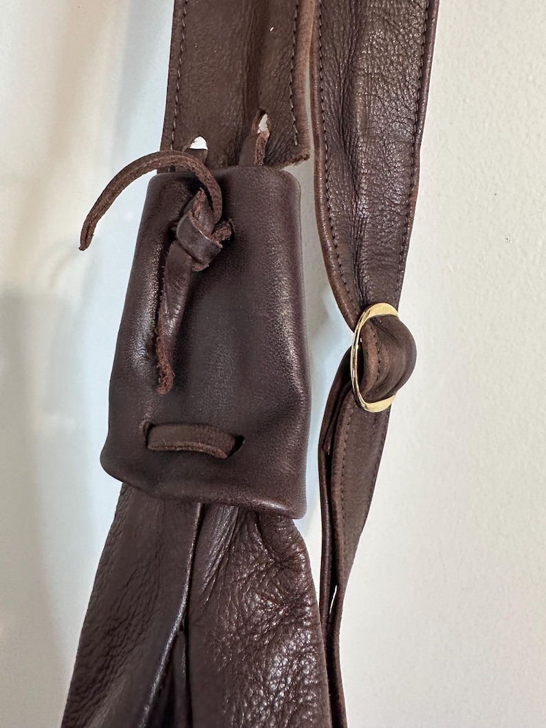 May include: Close-up of a dark brown leather bag with a drawstring closure. The bag features a long strap with a gold-colored metal ring. The leather has a textured appearance.