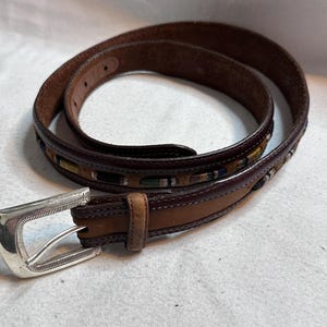 K&ouml;nnte beinhalten: Ein brauner Lederg&uuml;rtel mit silberner Schnalle. Der G&uuml;rtel hat ein dekoratives Webmuster in Gr&uuml;n-, Blau- und Gelbt&ouml;nen. Der G&uuml;rtel ist von mittlerer Breite und scheint ungetragen.