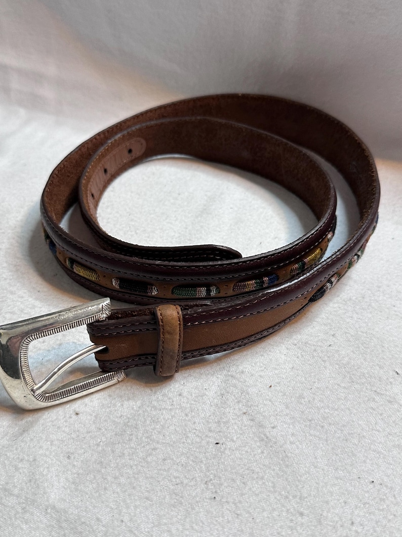 K&ouml;nnte beinhalten: Ein brauner Lederg&uuml;rtel mit silberner Schnalle. Der G&uuml;rtel hat ein dekoratives Webmuster in Gr&uuml;n-, Blau- und Gelbt&ouml;nen. Der G&uuml;rtel ist von mittlerer Breite und scheint ungetragen.