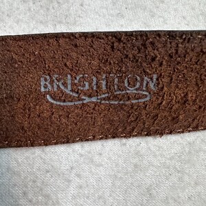 K&ouml;nnte beinhalten: Nahaufnahme eines braunen Lederg&uuml;rtels mit dem Markennamen "BRIGHTON" in Silber gepr&auml;gt. Der G&uuml;rtel hat eine strukturierte Oberfl&auml;che und einen gen&auml;hten Rand, was auf Haltbarkeit und Qualit&auml;t hindeutet. Das Bild konzentriert sich auf das Detail des G&uuml;rtels.