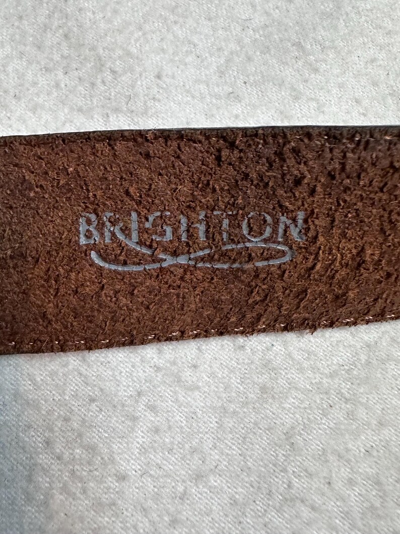 K&ouml;nnte beinhalten: Nahaufnahme eines braunen Lederg&uuml;rtels mit dem Markennamen "BRIGHTON" in Silber gepr&auml;gt. Der G&uuml;rtel hat eine strukturierte Oberfl&auml;che und einen gen&auml;hten Rand, was auf Haltbarkeit und Qualit&auml;t hindeutet. Das Bild konzentriert sich auf das Detail des G&uuml;rtels.