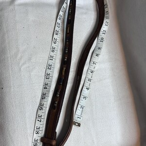 K&ouml;nnte beinhalten: Ein brauner Lederg&uuml;rtel mit dekorativen N&auml;hten und einer silberfarbenen Schnalle. Der G&uuml;rtel wird mit einem Ma&szlig;band gemessen, das eine L&auml;nge von ca. 102 cm anzeigt.