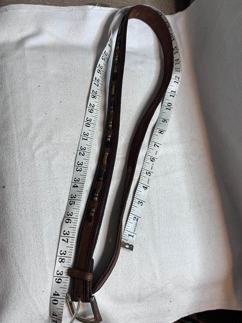 K&ouml;nnte beinhalten: Ein brauner Lederg&uuml;rtel mit dekorativen N&auml;hten und einer silberfarbenen Schnalle. Der G&uuml;rtel wird mit einem Ma&szlig;band gemessen, das eine L&auml;nge von ca. 102 cm anzeigt.