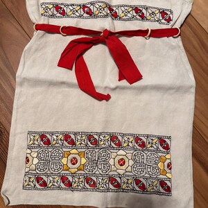 May include: A light-colored linen apron with embroidered floral patterns in red, yellow, and black. The apron has a red ribbon tie and two wooden rings. The embroidered design is at the top and bottom of the apron.