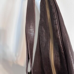 May include: A dark purple leather shoulder bag with a textured surface. The bag features a long strap, a gold-colored metal ring, and a zipper closure. The bag is designed with a rounded shape.