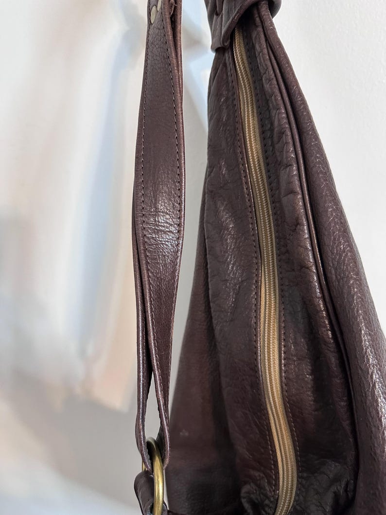 May include: A dark purple leather shoulder bag with a textured surface. The bag features a long strap, a gold-colored metal ring, and a zipper closure. The bag is designed with a rounded shape.