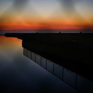 May include: A sunset sky with vibrant orange and deep blue hues reflects onto a calm body of water. A dark, silhouetted pier extends into the horizon, partially obscured by a blurred chain-link fence in the foreground, creating a stark contrast.