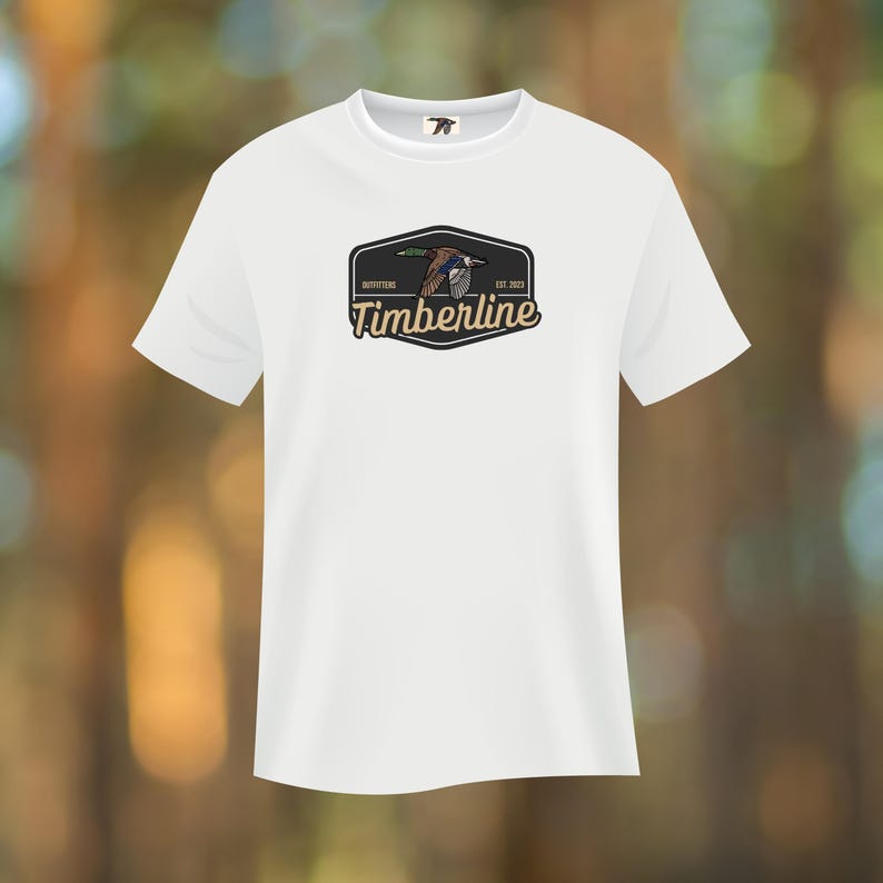 Puede incluir: Camiseta blanca con un gr&aacute;fico de un pato volando dentro de un emblema negro y verde. El emblema dice "Timberline Outfitters Est. 2023". La camiseta tiene cuello redondo y mangas cortas, adecuada para uso casual.