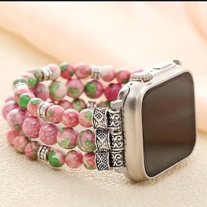 Op de afbeelding: Een Apple Watch-bandje met kralen met roze en groen gespikkelde kralen. De band heeft drie strengen ronde kralen met zilverkleurige metalen accenten en een zilveren horlogekast.