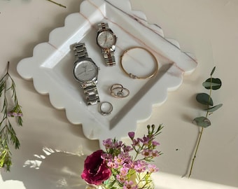 May include: A white marble tray with a scalloped edge holds two watches, a gold bracelet, and three rings. A bouquet of pink and white flowers sits below the tray, with greenery scattered around the scene.