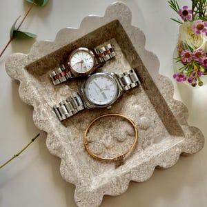 May include: A decorative, scalloped-edge tray in shades of beige and gray holds two wristwatches and a gold bracelet. One watch has a silver band and face, the other has a rose gold band and face. The bracelet is gold with a small charm.