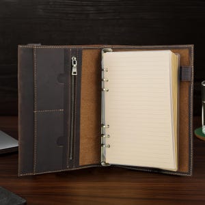 May include: Open brown leather journal with lined paper. The journal has a zipper closure, pockets, and a ring binder mechanism. The interior is a lighter brown color, and the journal is open, revealing the pages.