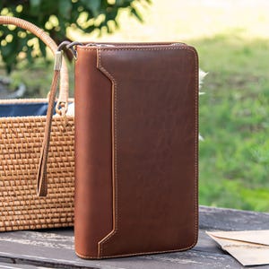 Puede incluir: Un portafolio de cuero marrón con cremallera y correa de muñeca. El diseño presenta un panel y costuras de color marrón claro. Una cesta de mimbre es visible en el fondo.