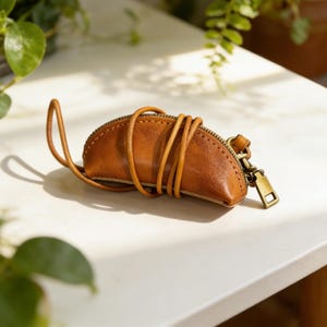 Puede incluir: Un estuche de cuero marrón con cierre de cremallera y una correa de cuero a juego. El estuche tiene forma de media luna y parece hecho a mano. El tirador de la cremallera y otros herrajes son de color latón. El estuche está sobre una superficie blanca.