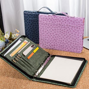 May include: Three document organisers in blue, purple, and green. The green organiser is open, revealing pockets, pens, a phone, and a notepad. The purple and blue organisers have a floral pattern and a handle. The organisers are on a woven surface.