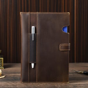 May include: A brown leather journal with a pen holder and a pen. The journal has a flap closure and a blue interior. The leather has a distressed look with visible stitching.
