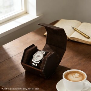 May include: A silver wristwatch with a white face is displayed inside a brown leather watch case. The case is open, revealing the watch against a soft, brown interior. A cup of coffee with latte art sits on a saucer nearby.
