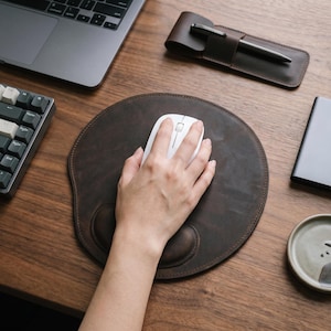 May include: A round dark brown leather mouse pad with a built-in wrist rest. The surface features visible stitching along the edges and a smooth matte texture. A white computer mouse is positioned on the centre of the pad, which is placed on a wooden desk.