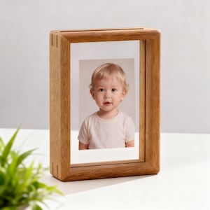 Puede incluir: Un marco de fotos rectangular de madera con una parte frontal de vidrio transparente. El marco contiene un retrato de un niño pequeño con una camiseta blanca. El marco es de madera marrón claro.