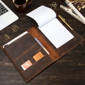 May include: A brown leather notebook cover with a notepad, pen, and card slots. The cover is open, showing a white notepad and a gold pen. A smartphone and two cards are visible in the pockets. A cup of coffee and a laptop are in the background.