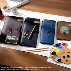 May include: Four leather-bound journals in brown, navy, and blue, each with a decorative charm and a brass-colored metal plate. A wooden palette with paint and two paintbrushes are also visible. The journals have solid-color covers and blank inner pages.