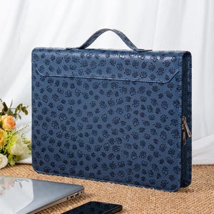 Pode incluir: Uma pasta de couro azul com uma alça e fecho de correr. A pasta é decorada com um padrão floral. Um laptop e um smartphone são visíveis em primeiro plano.