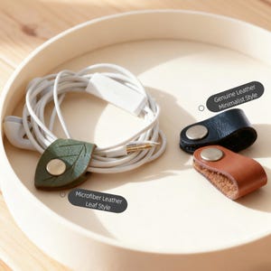 May include: Earbud cable organizers in a wooden tray. One is leaf-shaped, green, and microfiber leather. Two are minimalist style, black and brown, genuine leather. The text reads "Genuine Leather Minimalist Style" and "Microfiber Leather Leaf Style."