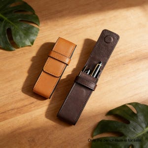 May include: Two leather pencil cases on a wooden surface. One is closed, tan-colored, and has a strap closure. The other is open, dark brown, and contains pens. The text at the bottom reads: Only the pencil case is for sale.