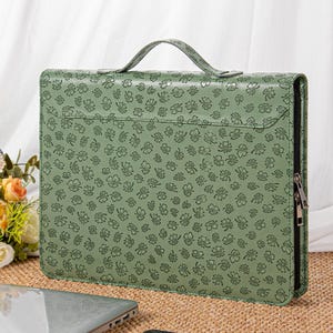 May include: A sage green briefcase with a handle and a zipper closure. The briefcase is covered in a pattern of small, dark green floral designs. The briefcase is sitting on a woven surface.
