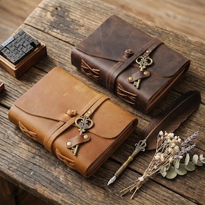 May include: Two leather-bound journals in dark brown and tan, each featuring a wrap-around strap secured with a brass-toned key charm. A wooden stamp set, a dark brown feather quill pen, and a small sprig of dried flowers are arranged nearby.