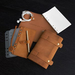 Könnte beinhalten: Eine Sammlung brauner Lederjournale, Notizbücher und Zubehör. Enthält ein Journal mit Stift, ein Notizbuch mit liniertem Papier und eine Ledertasche mit Riemen. Der Text "Customize your text or logo" ist sichtbar. Weiße Ohrhörer sind ebenfalls vorhanden.