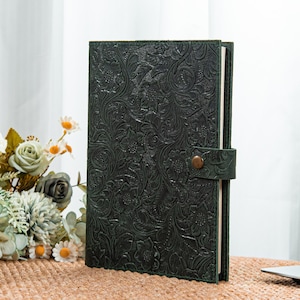 May include: Dark green journal with a floral embossed design. The journal has a strap closure with a bronze-colored button. The cover is a solid dark green color. The journal is open, revealing the white pages inside.