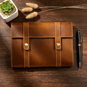May include: A brown leather portfolio with two vertical straps secured by brass-colored buttons. A black pen with gold accents rests beside the portfolio. A small succulent plant in a white pot and decorative dried grass are also present.