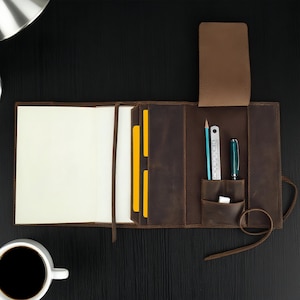 May include: A brown leather journal with a pen, pencil, and ruler in the pocket. The journal is open, revealing blank pages and yellow card holders. A cup of coffee sits to the side.