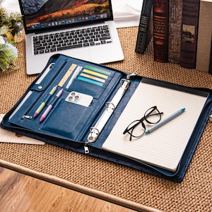 Może przedstawiać: Niebieskie skórzane portfolio z zapięciem na zamek błyskawiczny, otwarte, aby odsłonić notatnik, długopisy i smartfon. Portfolio ma również przegródki na karty i segregator z trzema pierścieniami. Laptop i książki są w tle.
