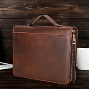 May include: A brown leather briefcase with a handle and a full-length zip. The briefcase has a distressed finish and visible stitching. The handle is stitched and reinforced. The briefcase is on a dark wooden surface.
