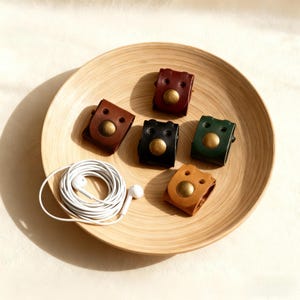 May include: A collection of six leather earbud organizers in various colors: brown, black, green, and tan. Each organizer features a brass button closure and is arranged on a wooden plate. A pair of white earbuds and cord are also present.