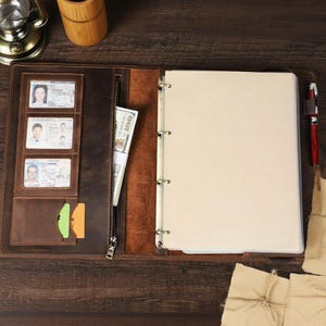 May include: Open brown leather binder with a zipper closure, multiple card slots, and a three-ring binder. The binder contains a stack of blank paper, a pen, and several identification cards.