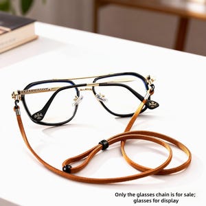 May include: Black and gold-rimmed eyeglasses with a brown leather glasses chain. The glasses have a double bridge and a decorative floral design on the temples. The chain is looped and has a black bead.