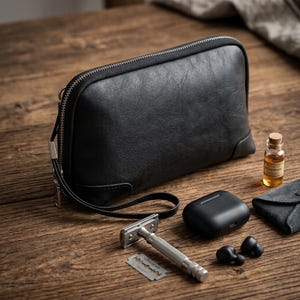 Könnte beinhalten: Eine schwarze Ledertasche mit Reißverschluss und Handschlaufe, ein silberner Sicherheitsrasierer aus Metall, eine Rasierklinge, ein schwarzes Ladeetui für kabellose Ohrhörer, zwei schwarze Ohrhörer und eine kleine Glasflasche mit Aftershave.