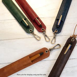 May include: An assortment of leather pen cases in various colors, including green, red, blue, brown, and tan. Each case holds a pen and has a metal clasp. The text at the bottom reads: "Pens are for display only, not for sale."