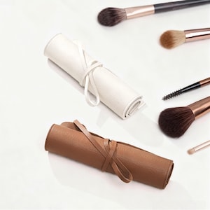 May include: A white and a brown leather makeup brush roll with tie closures. Several makeup brushes with brown and black handles and soft bristles are scattered around the rolls. The brushes are in various sizes and shapes.