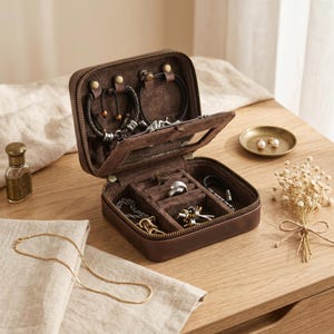 May include: A square, dark brown leather jewellery box with a zip closure. The interior is lined with dark brown fabric and features a top lid with hooks for necklaces and a bottom section with divided compartments for rings and bracelets.