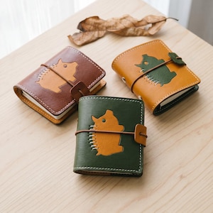Op de afbeelding: Drie kleine leren notitieboekjes met elastische sluitingen. Elke kaft is voorzien van een gestikt dierensilhouet in een contrasterende kleur. De notitieboekjes zijn bruin, geel en groen, met witte stiksels langs de dierenfiguren.