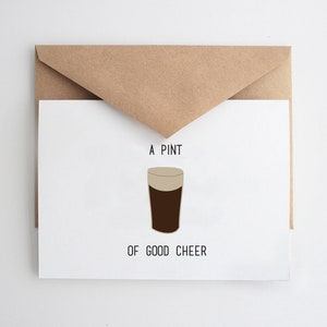 May include: A white card with a brown envelope on top. The card has black text that reads "A Pint of Good Cheer" and a cartoon illustration of a glass of dark beer.